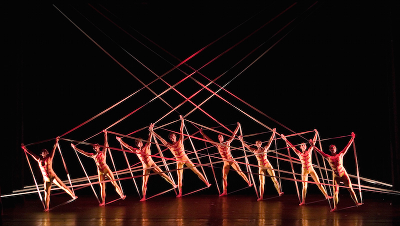 Dancers in unitards hold onto to tensile fabric with their hands and feet to create an intricate geometric pattern
